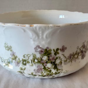 May include: White porcelain bowl with a scalloped rim and floral design. The bowl features a delicate pattern of green leaves and small purple and white flowers. The rim has a gold trim.