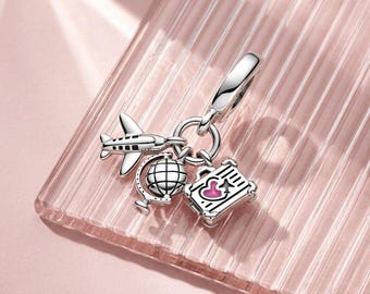 May include: A silver dangle charm featuring three distinct travel-themed pendants attached to a circular bail: a small airplane, a detailed globe on a stand, and a rectangular suitcase with a pink heart and a small airplane icon on its front surface.