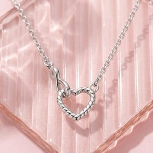 May include: A silver-toned necklace featuring a delicate chain and a pendant composed of an infinity symbol linked to a heart-shaped charm. The heart charm has a twisted rope-like texture, and the entire piece is crafted from polished metal.