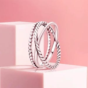 May include: A silver ring with three overlapping bands. One band has a beaded pattern, the others a twisted rope design. The ring is presented on a pink surface, set against a soft pink background.