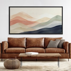 Puede incluir: Una gran obra de arte abstracta enmarcada que representa montañas de acuarela en capas en tonos coral, beige y azul. La obra de arte cuelga sobre un sofá de cuero marrón con cojines. Una mesa de centro de madera y una cesta tejida completan la escena.