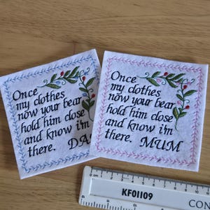 May include: Two white felt squares with embroidered floral designs and text. The top square has pink stitching and the text "Once my clothes now your bear hold him close and know I'm there. MUM". The bottom square has blue stitching and the text "Once my clothes now your bear hold him close and know I'm there. DA".