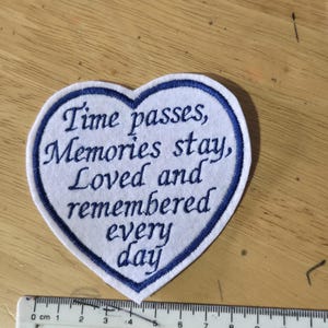 May include: A heart-shaped white fabric patch with a blue embroidered border and the blue text "Time passes, Memories stay, Loved and remembered every day". A ruler is positioned at the bottom, showing the patch is approximately 10 cm wide.