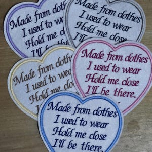 May include: Four heart-shaped fabric patches with embroidered text. The text reads "Made from clothes I used to wear Hold me close I'll be there." The patches are in different colours: purple, grey, yellow, and blue. The blue patch has "XX" embroidered at the bottom.