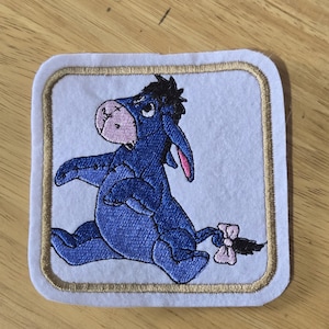 May include: A square fabric patch with rounded corners featuring an embroidered blue donkey character with a light pink muzzle and a small pink bow on its tail. The design is set against a white background and framed by a gold-coloured stitched border.