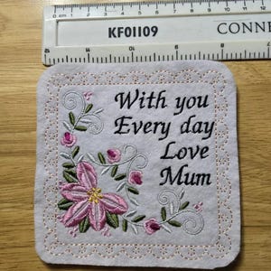 May include: White felt square with pink and green embroidered flowers and the text "With you Every day Love Mum".