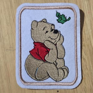May include: Embroidered patch showing a seated cartoon bear in a red shirt, gazing at a green leaf. The bear is tan with black details, set against a pale blue background with a beige border. The patch is rectangular with rounded corners.