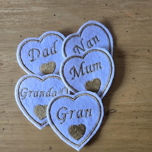May include: Five white felt heart-shaped patches with gold embroidered text. The text reads "Dad", "Nan", "Mum", "Grandad", and "Gran". Each heart has a gold heart-shaped accent. Ideal for crafting or gifts.