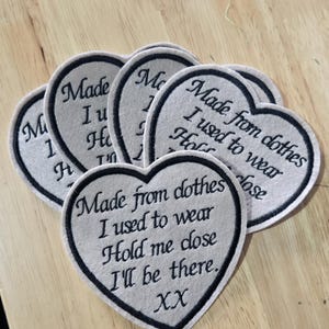 May include: A collection of heart-shaped fabric patches in a light grey colour with black embroidered borders. Each patch features the black embroidered text: Made from clothes, I used to wear, Hold me close, I'll be there, XX.