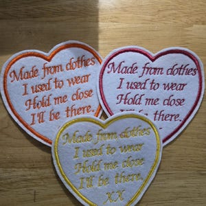 May include: Three heart-shaped embroidered patches. The patches are white with orange, red, and gold borders. Each patch features the embroidered text: "Made from clothes I used to wear, Hold me close, I'll be there."