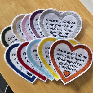 May include: A collection of heart-shaped fabric patches with embroidered borders in various colours including silver, purple, pink, gold, blue, red, light blue, yellow, and orange. Each patch features the black embroidered text: Once my clothes now your bear. Hold me close and know I'm there.