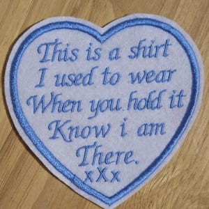 May include: A white heart-shaped patch with blue stitching around the edges. The patch has the text "This is a shirt I used to wear When you hold it Know I am There. XXX" embroidered in blue thread.