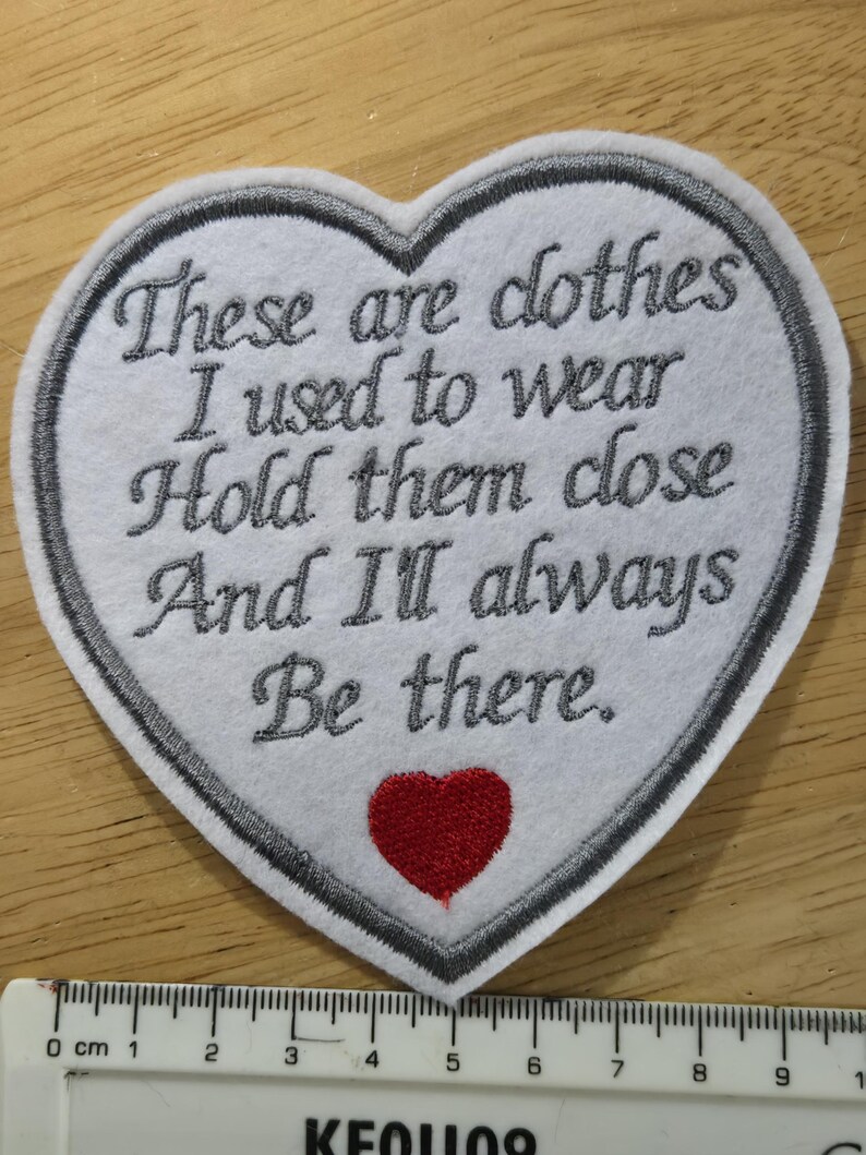 Puede incluir: Un parche en forma de coraz&oacute;n con texto bordado: "These are clothes I used to wear Hold them close And I'll always Be there." El parche tiene un fondo blanco, un borde gris y un coraz&oacute;n rojo. Una regla muestra las medidas en cent&iacute;metros.