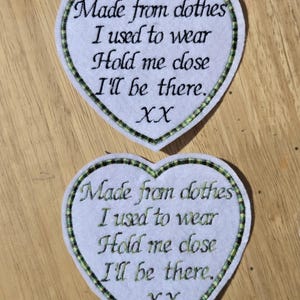 May include: Two white heart-shaped patches with green and black trim. Each patch has embroidered text: "Made from clothes I used to wear, Hold me close, I'll be there. XX". The patches are on a wooden surface.