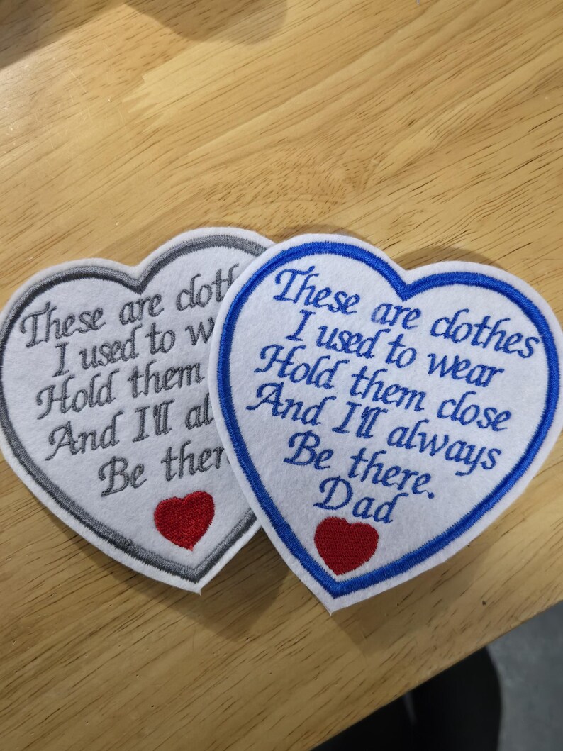 Puede incluir: Dos parches bordados en forma de coraz&oacute;n. Uno tiene un borde gris, el otro un borde azul. Ambos tienen el texto "These are clothes I used to wear Hold them close And I'll always Be there." y un coraz&oacute;n rojo.