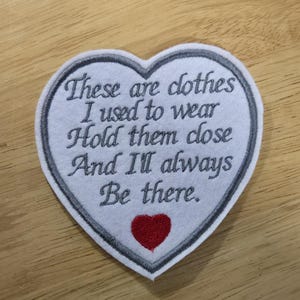 Puede incluir: Un parche en forma de coraz&oacute;n con texto bordado: "Esta es la ropa que sol&iacute;a usar. Abr&aacute;zala y siempre estar&eacute; ah&iacute;". El parche tiene un fondo blanco, un borde gris y un coraz&oacute;n rojo.