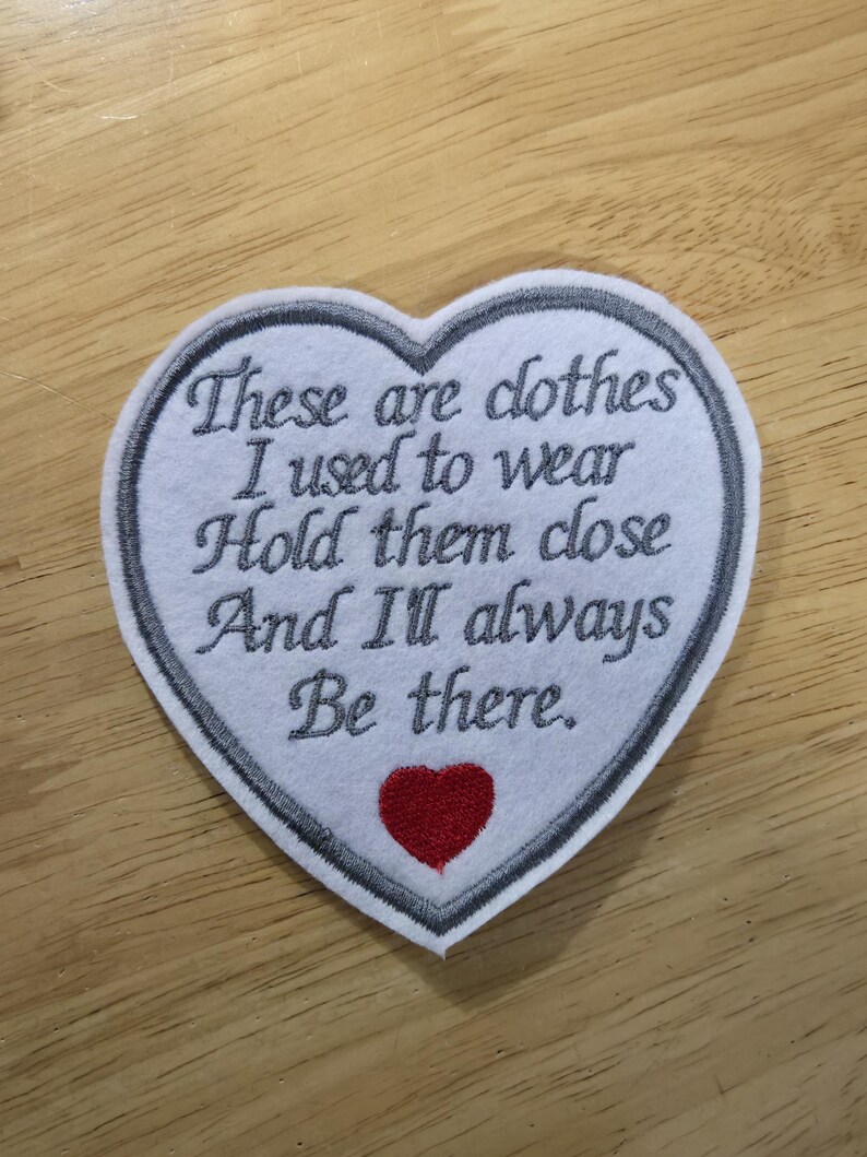Puede incluir: Un parche en forma de coraz&oacute;n con texto bordado: "Esta es la ropa que sol&iacute;a usar. Abr&aacute;zala y siempre estar&eacute; ah&iacute;". El parche tiene un fondo blanco, un borde gris y un coraz&oacute;n rojo.
