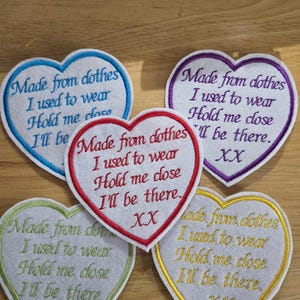 May include: Five heart-shaped embroidered patches with the text: "Made from clothes I used to wear Hold me close I'll be there. XX." Each patch has a white centre and a coloured border: blue, purple, red, green, and gold.