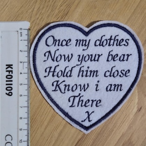 May include: White heart-shaped felt patch with blue embroidered edging. The patch has the text "Once my clothes Now your bear Hold him close Know I am There X" embroidered in black.