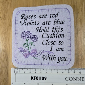 May include: A white felt cushion with purple embroidery that reads "Roses are red, Violets are blue, Hold this Cushion Close so I am With you". The embroidery also includes a purple flower with a green stem and a purple bow.