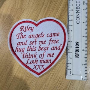 May include: White felt heart-shaped patch with red embroidered edges. The text "Riley The angels came and set me free hug this bear and think of me Love mam XXX" is embroidered in red.