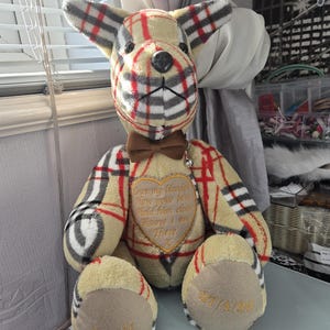 May include: A plush teddy bear crafted from beige, black, and red plaid fabric. It features a brown bow tie, a heart-shaped patch with the embroidered text 'Once my blanket, Now your bear, Hold him close, Know I am There x', and feet with 'Ernie' and '27/4/26' embroidered in gold.