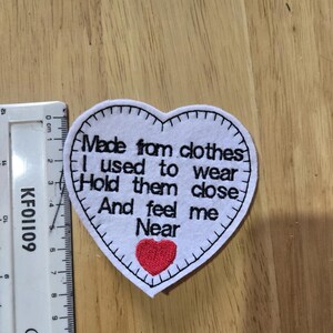 May include: A white heart-shaped patch with black stitching and the text "Made from clothes I used to wear Hold them close And feel me Near". A small red heart is at the bottom.