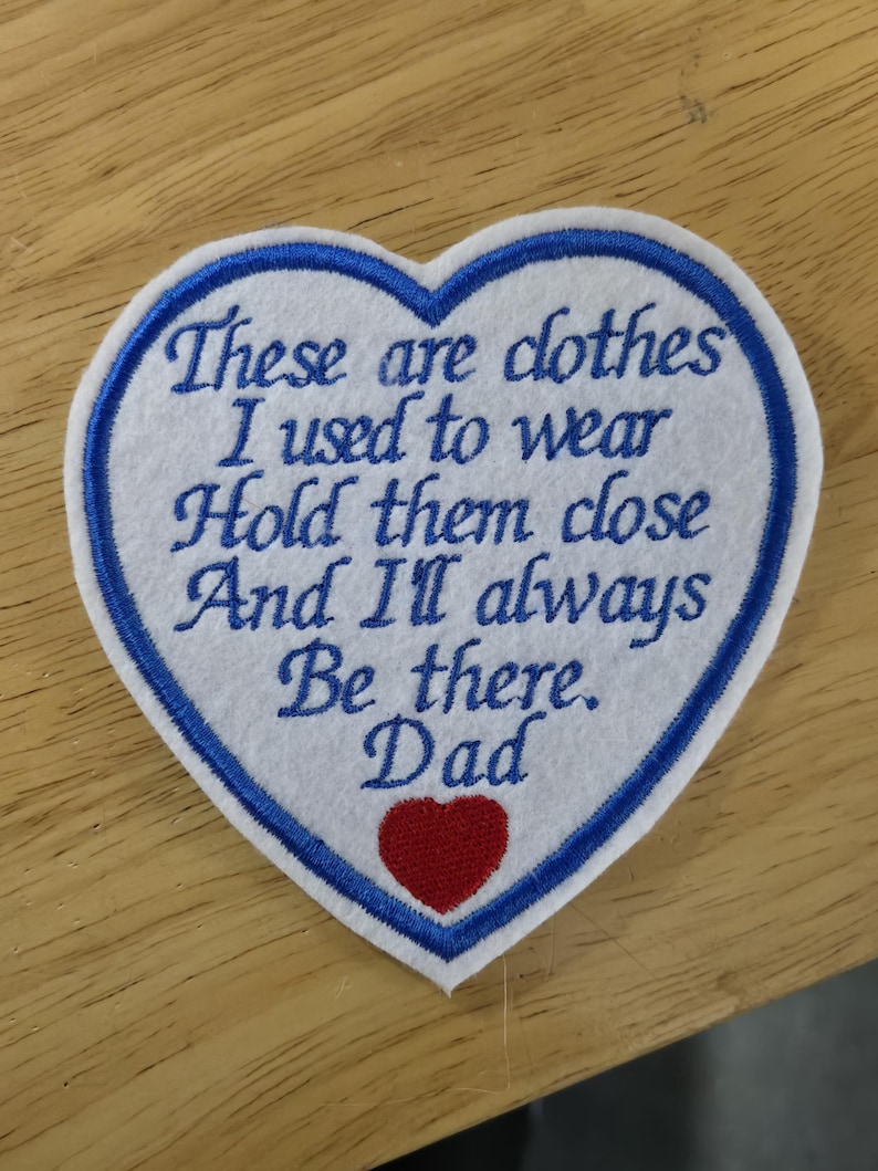 Puede incluir: Un parche en forma de coraz&oacute;n con un centro blanco y un borde azul. El parche presenta texto bordado en azul, que dice: "These are clothes I used to wear Hold them close And I'll always Be there. Dad." Un coraz&oacute;n rojo en la parte inferior.