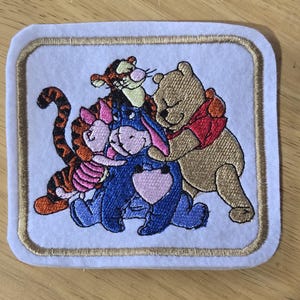 May include: A square embroidered patch on a white felt background featuring a group of four cartoon characters: a yellow bear in a red shirt, a blue donkey, a pink piglet, and an orange striped tiger, all enclosed by a gold stitched border.