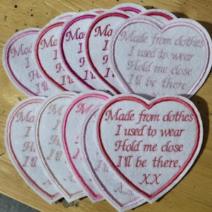 May include: A group of heart-shaped patches with embroidered text. The patches are white with pink and red stitching. The text reads "Made from clothes I used to wear Hold me close I'll be there."