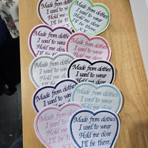 May include: A collection of heart-shaped fabric patches with embroidered text reading: Made from clothes I used to wear, Hold me close, I'll be there, XX. The patches feature various coloured borders including brown, green, pink, black, purple, blue, and light blue.