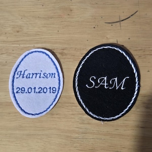 May include: Two oval-shaped fabric patches with embroidered borders. The white patch features blue embroidery reading Harrison and 29.01.2019. The black patch features white embroidery reading SAM. Both patches have a decorative stitched edge.
