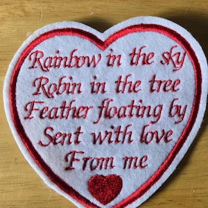May include: A white felt heart-shaped patch with red embroidered trim and text. The text reads "Rainbow in the sky, Robin in the tree, Feather floating by, Sent with love, From me." A small red heart is at the bottom.