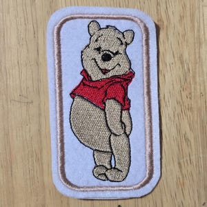 May include: Embroidered patch of a smiling bear in a red shirt. The bear is tan with black features, set against a light blue background with a brown border. The patch is rectangular with rounded corners.