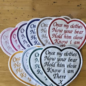 May include: A collection of heart-shaped fabric patches with embroidered borders in various colours. Each patch features the text: Once my clothes, Now your bear, Hold him close, Know i am There, followed by an X. The patches are stacked in a fan shape.