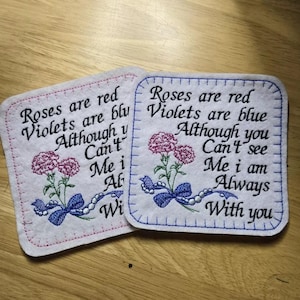 May include: Two embroidered felt coasters with the text "Roses are red, Violets are blue, Although you can't see me, I am always with you". The coasters are pink and blue with a flower design.