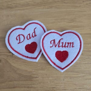 May include: Two white heart-shaped patches with red embroidered outlines and a small red heart in the centre. One patch says "Dad" in red script, and the other says "Mum".