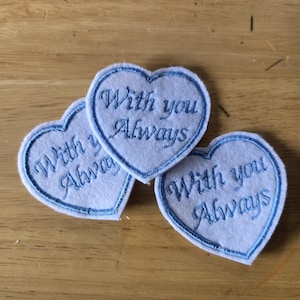 May include: Three white felt heart-shaped patches with blue embroidered borders. Each patch has the phrase "With you Always" embroidered in blue script. The patches are arranged on a light brown wooden surface.