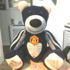 May include: A blue and white teddy bear wearing a Manchester United jersey with an Adidas logo on each foot. The bear has a heart-shaped patch with the text "This is a jacket I used to wear when you hold it know I am there Love Claire Z".