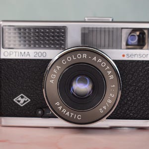 Könnte beinhalten: Eine Vintage Optima 200 Kamera mit einem schwarzen, strukturierten Gehäuse und silbernen Akzenten. Das Objektiv ist mit "AGFA COLOR-APOTAR 1:2.8/42" beschriftet. Das Wort "sensor" ist über dem Objektiv aufgedruckt. Eine klassische Kamera.