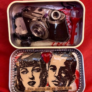 May include: A vintage tin box with a silver trim and a red heart. The box is open and inside there are two black and white images of a man and a woman. The text "Then suddenly she was back in his" is visible at the bottom of the box.