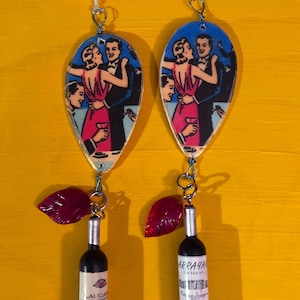 May include: A pair of dangle earrings featuring a vintage-style illustration of a couple dancing, with a wine bottle charm hanging below. The wine bottle charm has a red glass leaf and a silver top.