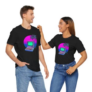 Peut inclure: T-shirts noirs avec un motif d'ordinateur rétro, un palmier et un coucher de soleil rose, violet et bleu. Un homme et une femme portent ces t-shirts avec des jeans bleus.