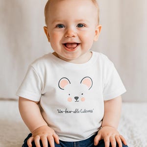 May include: A baby wearing a white t-shirt with a bear face design and the words "Un-bear-able Cuteness". The baby is also wearing blue denim jeans. The background is a soft, neutral colour.