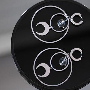 May include: Silver earrings with a crescent moon, a small clear bead, and a circular hoop. The earrings are displayed on a black, reflective surface. The design includes two large circles and two smaller circles.