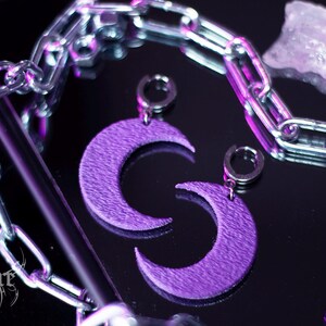 May include: A pair of purple crescent moon earrings with silver hoops. The earrings are laying on a black surface with a silver chain and a clear crystal.