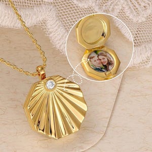 Op de afbeelding: Goudkleurige medaillonketting met een geometrische, waaiervormige hanger. De hanger kan worden geopend om een kleine foto te onthullen. De ketting heeft een fijne ketting en een kleine, heldere edelsteen.