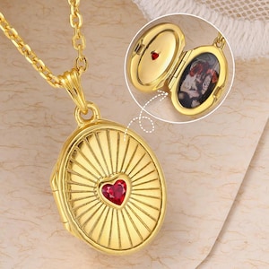 Könnte beinhalten: Goldfarbene Halskette mit einem ovalen Medaillon, das einen roten herzförmigen Edelstein enthält. Das Medaillon öffnet sich und zeigt ein kleines Foto. Die Kette ist filigran und goldfarben. Ein schönes Schmuckstück.