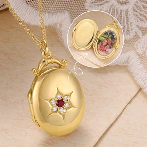 May include: A gold oval locket pendant on a matching gold chain. The front of the locket features a star-shaped engraving set with a central red gemstone surrounded by six small clear stones. An inset shows the locket open with a photograph inside.