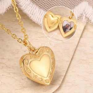 Op de afbeelding: Een goudkleurige hartvormige medaillonketting. Het medaillon is in close-up geopend en toont een kleine foto. De hartvormige hanger is versierd met delicate bloemen- en sterpatronen. De ketting is ook goudkleurig.